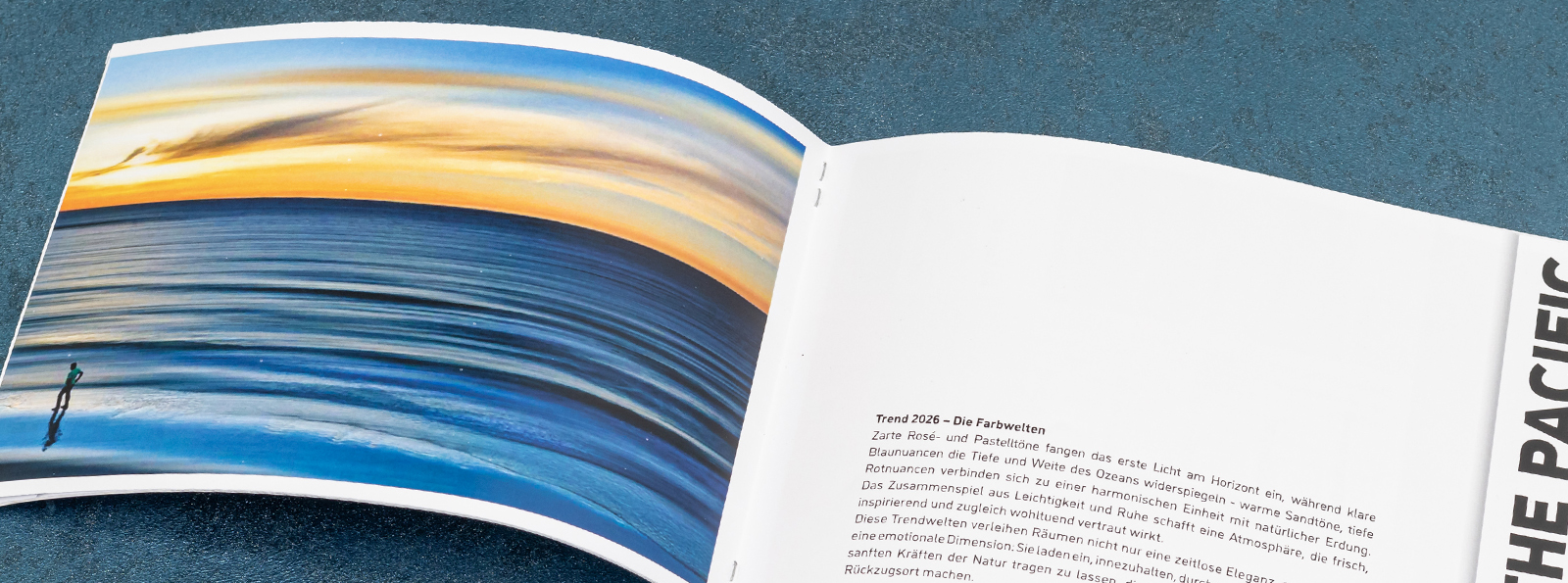 Ein geöffnetes Buch liegt auf einem blauen Untergrund. Die linke Seite zeigt ein Bild mit sanften Wellen und einem Sonnenuntergang. Die rechte Seite enthält Text in deutscher Sprache, der mit "THE PACIFIC" überschrieben ist.